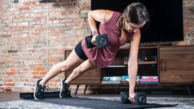 A woman does pushups using dumbbells on an exercise mat.