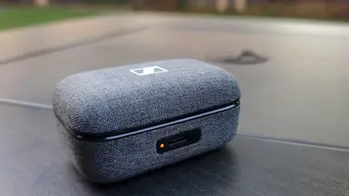 A gray fabric earbuds case sits on a black table with an orange LED at the front.