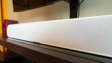 A white and tubular soundbar sits on a brown and black TV console with a TV mounted above.