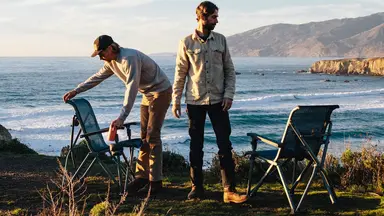 Two travelers setting their beach chairs up beside a large body of water.