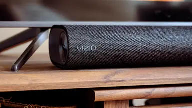 A fabric covered black Vizio soundbar sits below a TV on a wooden console.