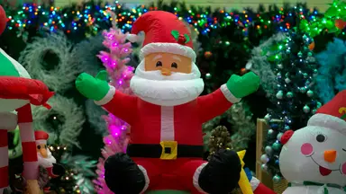 A light-up inflatable Santa decoration surrounded by other Christmas iconography: trees, colorful Christmas lights, an inflatable snowman, white wreaths, and more.