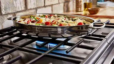 Close-up of a flavorful pasta dish in a silver pain. It sits on a gas stovetop with a blue flame glowing beneath it.