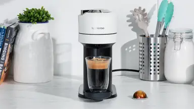 Nespresso Vertuo coffee machine on marble countertop in the center of books, potted plants, utensils and sugar.