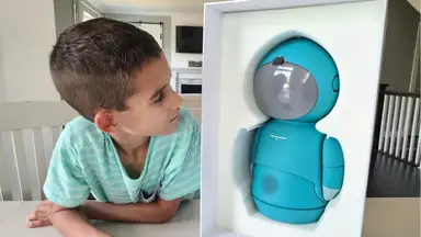 Small child smiling next to the Embodied Moxie robot in kitchen setting at table.