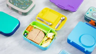 Bento boxes, cheese stick, carrots, apple, a sandwich, and M&Ms in a lunchbox, laid out on a marble counter.