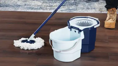 Person using Casabella Clean Water Spin Mop to clean hardwood floor surface next to bucket inside of modern living room setting.
