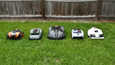 Five robot lawn mowers lined up together in the green grass with a fence behind