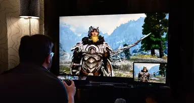 A man sits in a dark room playing a video game with a Viking-like character on the screen