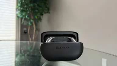An open black charging case with hearing aids inside sits on top of a glass table with a tree in the background