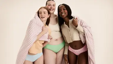 Three smiling women of diverse body types wearing period underwear and wrapped in a pink-striped blanket, posing together confidently.