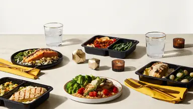 Multiple meals with seared chicken, salmon, vegetables on a beige table with geometric gold objects interspersed