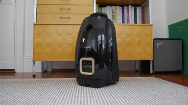 Black Miele Guard L1 Cat & Dog Vacuum sitting on a striped grey and white rug in front of tan drawers and a shelf.