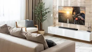 A bright, modern living room featuring an LG Dolby Atmos soundbar mounted beneath a large flat-screen TV on a light wood accent wall. The minimalist space includes a beige sectional sofa, white media console, indoor plant, and soft neutral decor, showcasing a clean and immersive home entertainment setup.