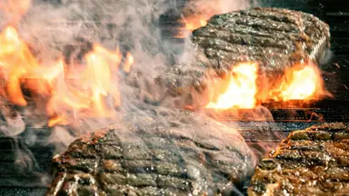 Steaks grilling over open flames with smoke rising.