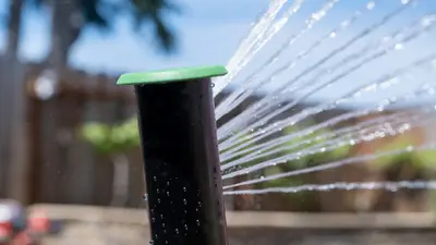 Irrigreen Smart Sprinkler 3 spraying water to the right, outside in front of a blue sky, tree, and building.