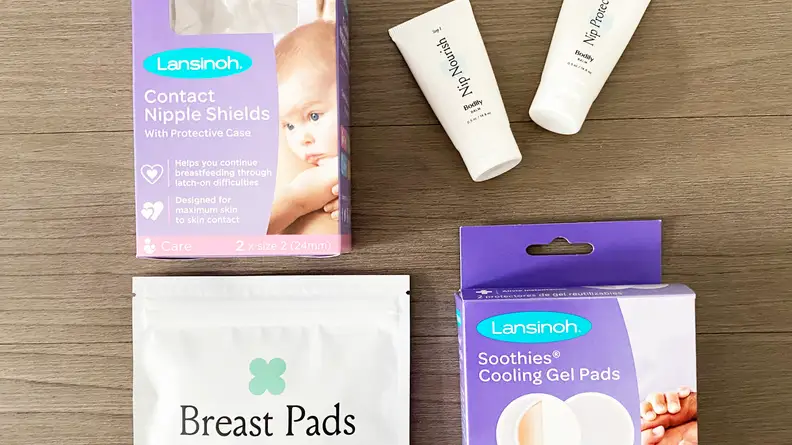 A set of breastfeeding accessories is laid out for display: Lansinoh contact nipple shields, Nip Nourish bodily balm, breast pads, and Lansinoh Soothies cooling gel pads.