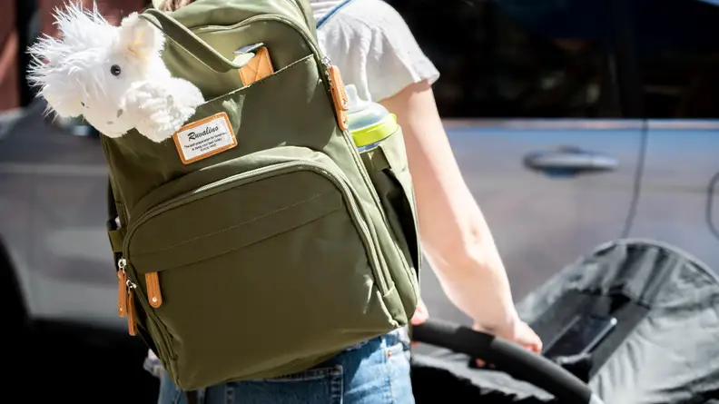 A sage green Ruvalino backpack-style diaper bag on the shoulder of a parent pushing a stroller.