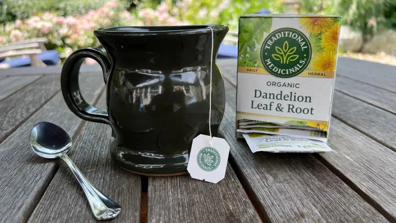 A mug with a tea bag string sit on an outdoor table between a spoon and a box of dandelion leaf and root tea.