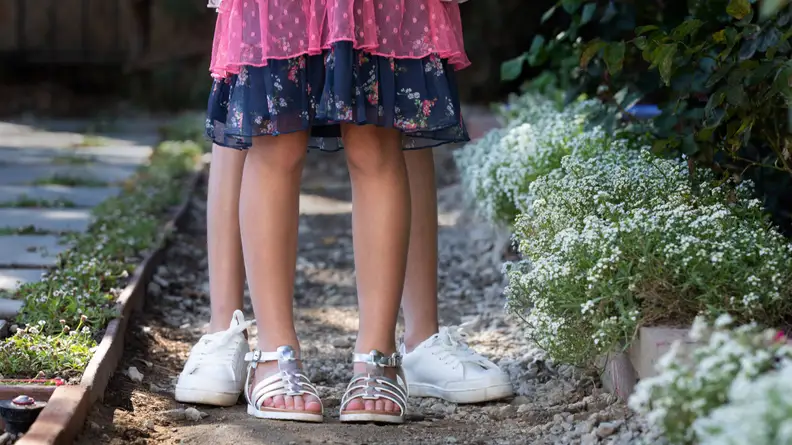 A pair of children's feet wearing silver sandals in front of a pair of children's feet wearing white tennis shoes.
