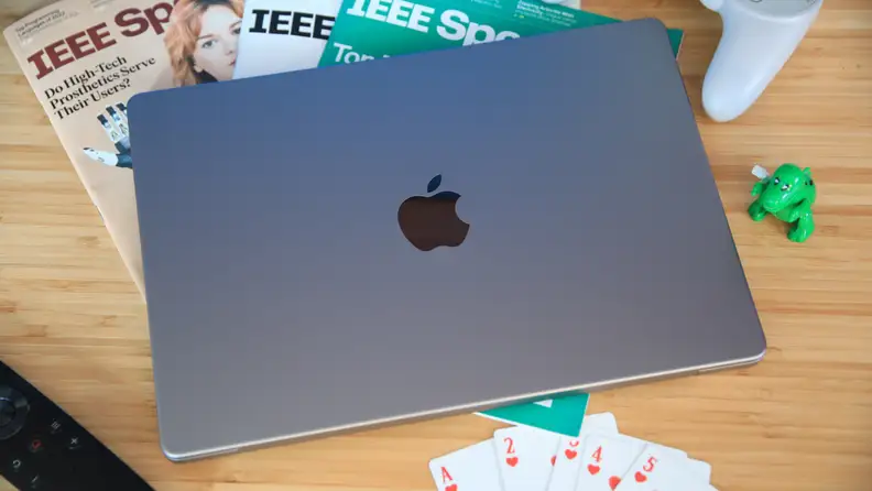 The Apple MacBook Pro 14 (2023) sitting on a wood table, closed, next to playing cards, magazines, and a green, toy dinosaur.