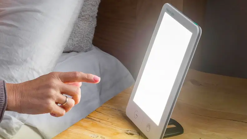 A close-up of the Happy Light in a bedroom setting. A hand reaches out to adjust it.