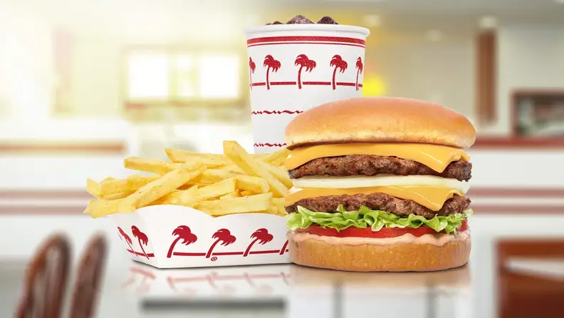An In-N-Out double cheeseburger with fries and a drink with blurred image of dining area as background