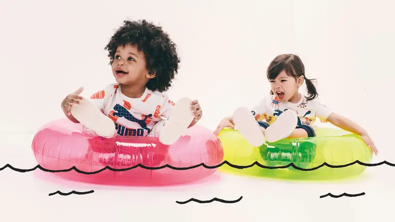 Two toddlers sit on tubes on a cartoon lake, the left toddler is a Black baby girl wearing white sneakers and a white tshirt, and the right toddler is a white girl with brown big tails