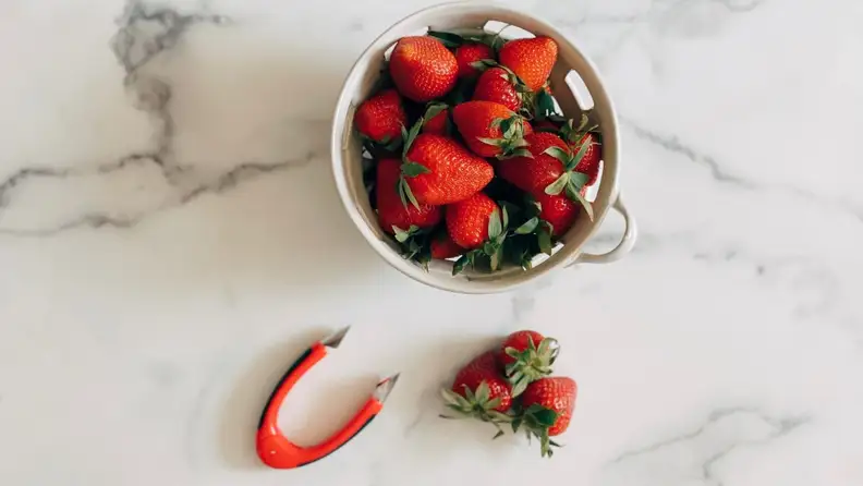 A bowl of strawberries next to the red OXO strawberry huller on a marble surface.