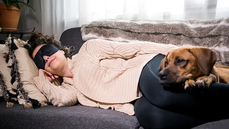 A person laying on the couch wearing a sleep mask with their dog.