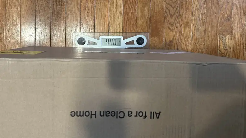 Cardboard box sitting on a clear scale over a hardwood floor, showing 44 pounds.
