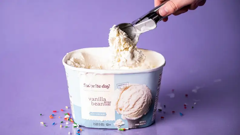 A hand scooping into a tub of vanilla ice cream on a purple background, surrounded by rainbow sprinkles.