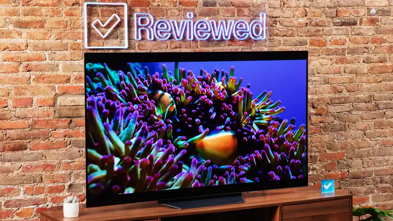 The LG B2 on a wooden table in front of a brick wall with a neon Reviewed sign displaying an ocean scene.