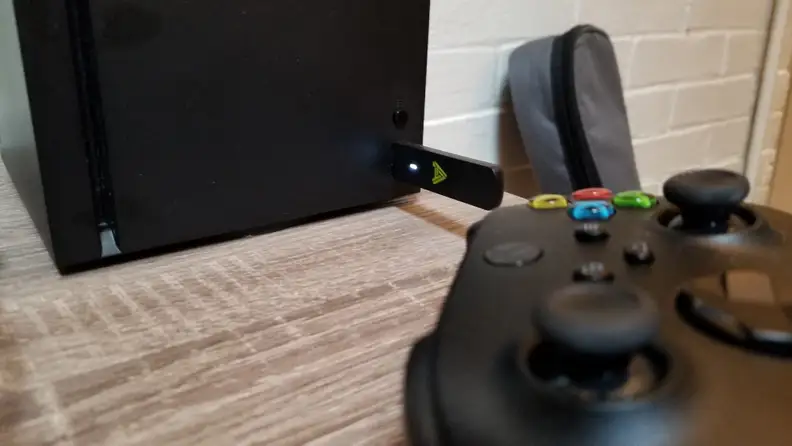 A USB dongle is inserted into the front of an Xbox Series X console. In the foreground, out of focus, is an Xbox controller.