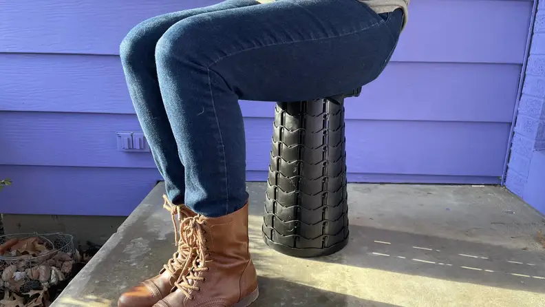 A person sitting on the Giraffe Creation Telescopic Stool outside, with a purple background.