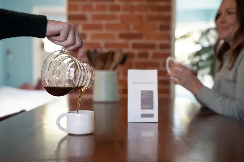 Coffee poured from a glass server into a mug beside a Wonderstate bag and a smiling woman