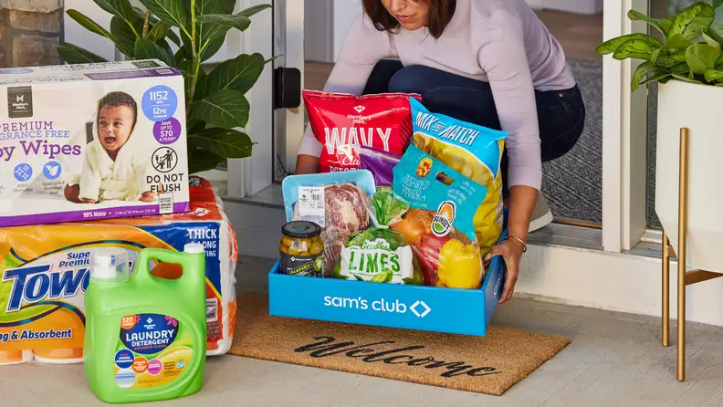 A woman brings a Sam’s Club grocery box filled with fresh produce and pantry staples inside her home, surrounded by bulk household items on the doorstep.