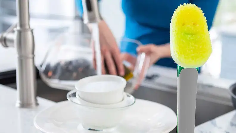 Standing Scrub Daddy Soap Wand in front of cleaned plates, a sink and someone washing a pan in a kitchen.