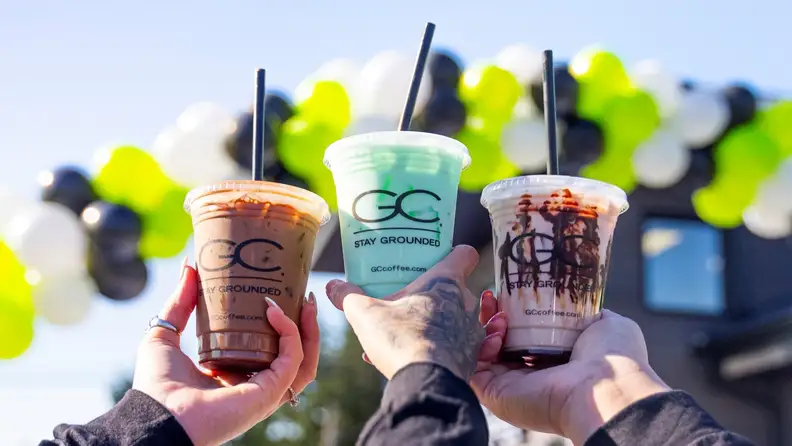 Three hands holding up iced drinks in GC Coffee cups against a balloon arch