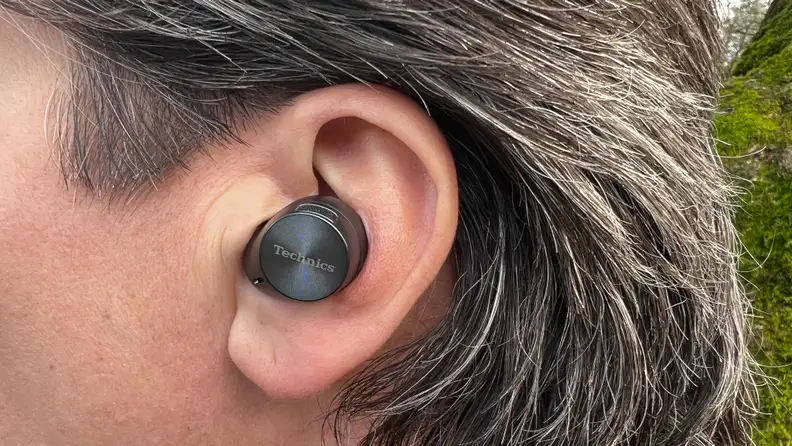 The gunsmoke colored wireless earbud sits in an ear of a person with salt and pepper brown hair.