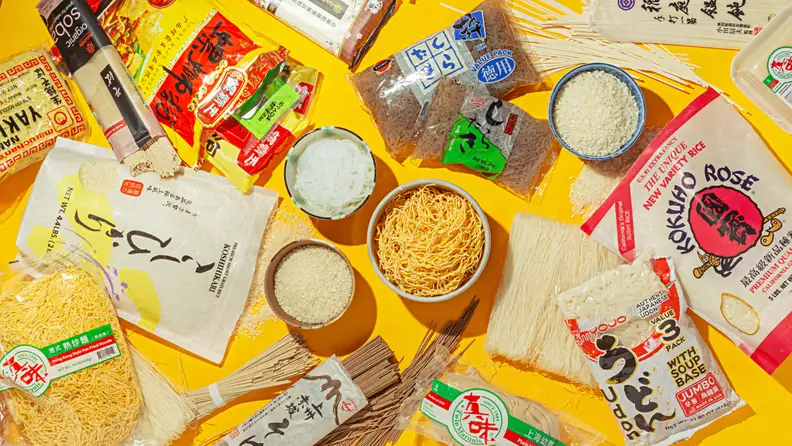 On a yellow table, there are various types of instant ramen, luosifen, udon noodles, white rice, and more scattered around.