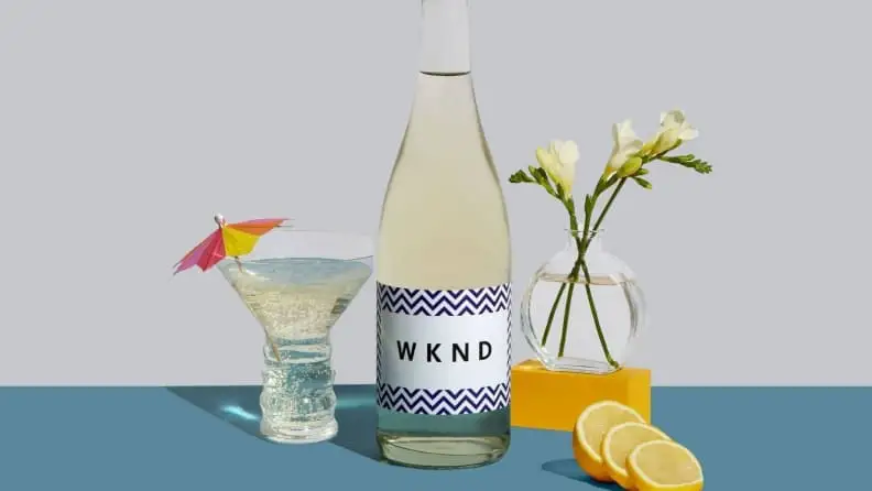 An artistic image featuring a bottle of white wine, a wine glass filled with the white wine and topped with a decorative umbrella, plus some lemon sliced and a small vase with flowers on the side.