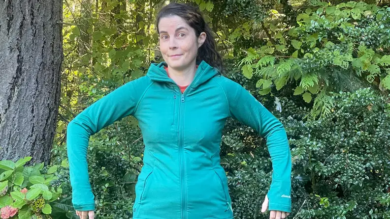 A woman outside next to trees, wearing a blue Eddie Bauer Women's High Route Full-Zip Grid Fleece Hoodie.
