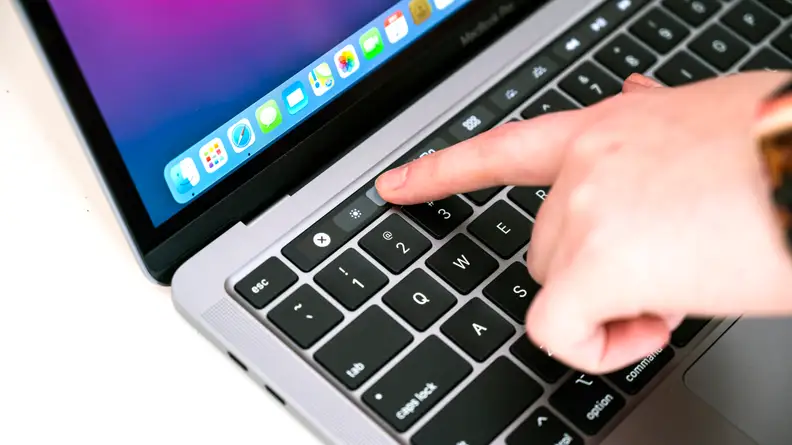 Close-up: A fingertip presses the brightness control on a MacBook Pro's touchscreen toolbar (at the top of the keyboard).