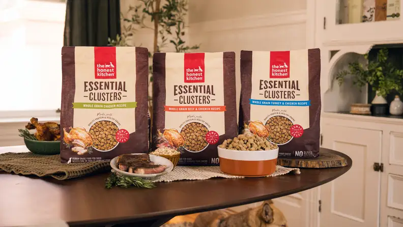Three bags of The Honest Kitchen dog food Essential Clusters in chicken, beef & chicken, and turkey & chicken recipes displayed on a table with fresh ingredients.