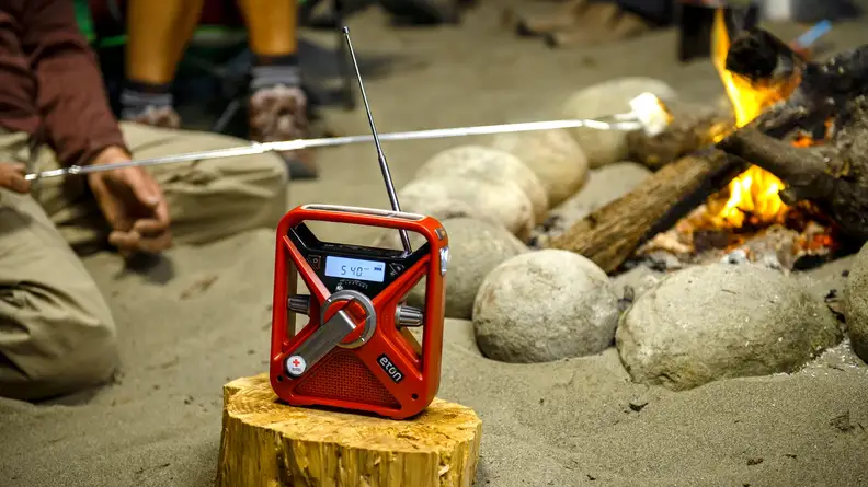 Red Etón FRX3+ Multi-Powered Weather Alert Radio sitting on top of wooden log next to person roasting marshmallow on campfire.