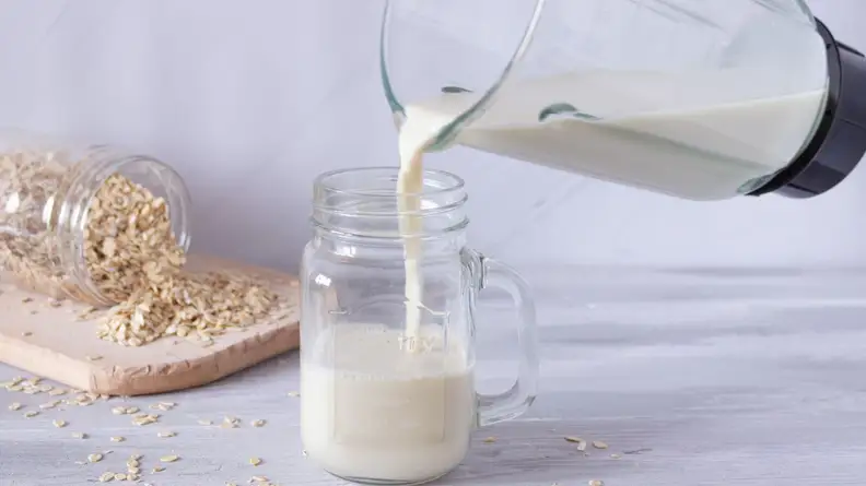 Oat milk from blender being poured into mason jar next to spilled mason jar of oats on top of wooden cutting board.
