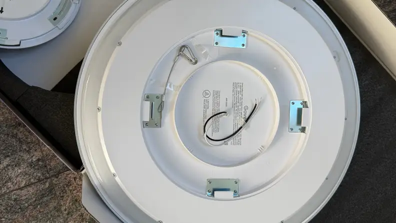 Rear view of a round flush-mount smart ceiling light showing integrated LED housing, electrical wiring leads, safety cable, and metal mounting brackets used during installation.