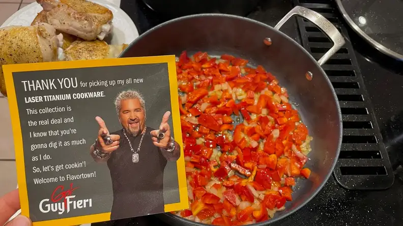 Person holding up a thank you note from Guy Fieiri in front of a skillet filled with chopped up peppers next to a cooked plate of chicken thighs.