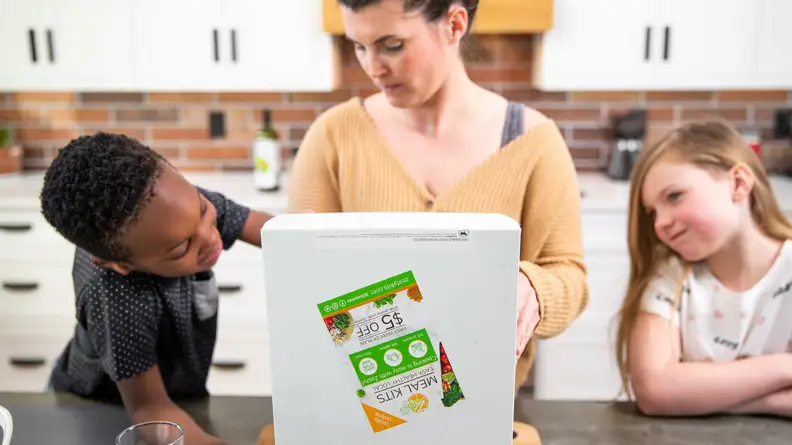 A woman opening the Zesty Kits meal kit with two children watching.